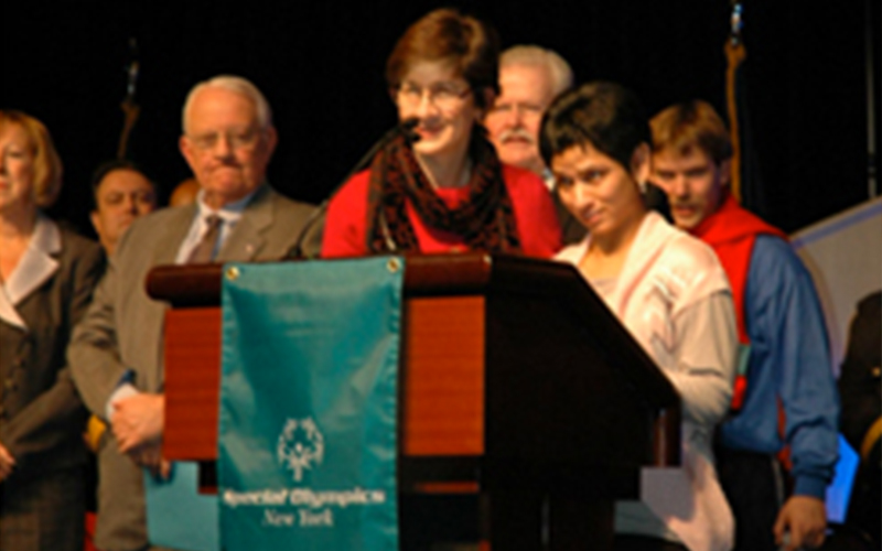 Ann Costello peaking at a podium at the Special Olympics Winter Games with a group of people behind her.