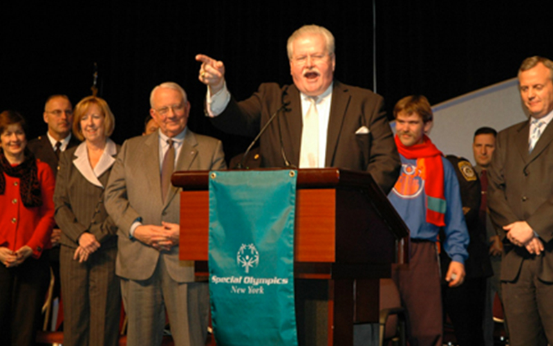 Man speaking at a podium at the Special Olympics Winter Games with a group of people behind him.